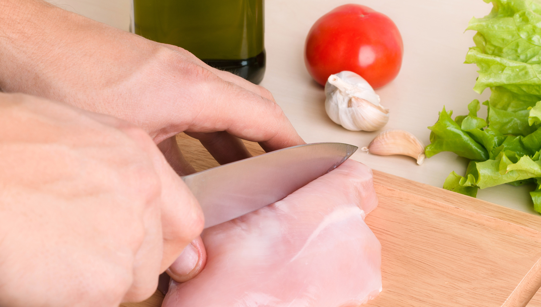 Das Bild zeigt eine Hand, die Hähnchenfleisch mit einem Messer zerschneidet.