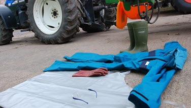 Das Bild zeigt Schutzkleidung wie Overall, Handschuhe, Schürze, Schutzbrille und Gummistiefel vor einem Traktor. (Quelle: Dittmar / SVLFG)